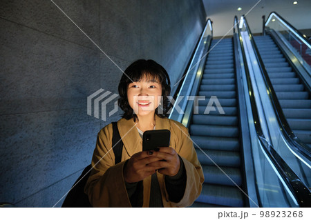 Cute young woman going down the escalator to the tube, using subway metro to commute to work or university, standing with smartphone 98923268