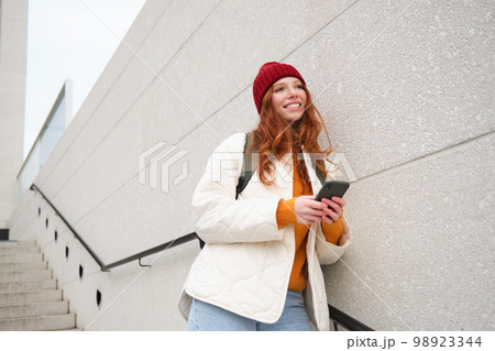 Happy girl student in red hat, holds smartphone, tourist looks at map app on her phone, explores sightseeing, texts message, looks for couchsurfing, rents place to stay online 98923344