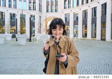 Technology, application and cellular concept. Smiling asia woman uses mobile phone, holds smartphone and reads text message, walks on streets of city centre 98923385
