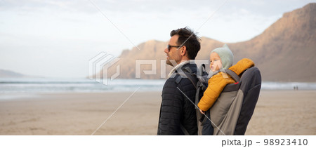 Father rising hands to the sky while enjoying pure nature carrying his infant baby boy son in backpack on windy sandy beach of Famara, Lanzarote island, Spain at sunset. Family travel concept Father rising hands to the sky while enjoying pure nature carrying his infant baby boy son in backpack on windy sandy beach of Famara, Lanzarote island, Spain at sunset. Family travel concept 98923410