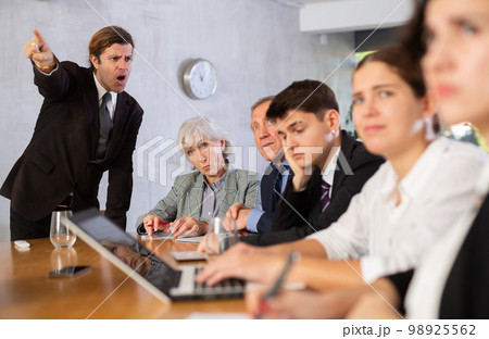 Displeased male manager scolding at young, adult, mature office workers sitting with reports and laptops at table in meeting room 98925562