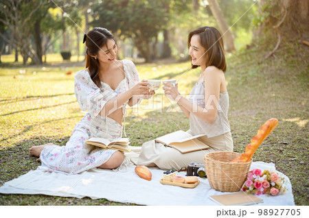 Two attractive Asian women are enjoying an afternoon tea picnic in the beautiful park, sipping tea 98927705