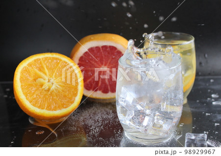 splashes of water over a glass of water and ice. Nearby are fruits orange and grapefruit, ice. On a black background. Refreshing cold ice drink splashes of water over a glass of water and ice. Nearby are fruits orange and grapefruit, ice. On a black background. Refreshing cold ice drink 98929967