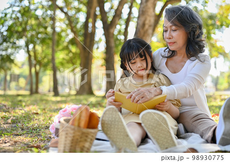 Happy Asian grandmother reading fairly tales to her cute granddaughter while relaxing in the park. Happy Asian grandmother reading fairly tales to her cute granddaughter while relaxing in the park. 98930775