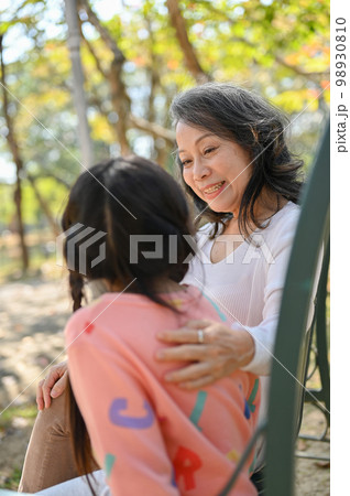 Caring Asian grandmother tells a story to her granddaughter while relax on a bench in the park Caring Asian grandmother tells a story to her granddaughter while relax on a bench in the park 98930810