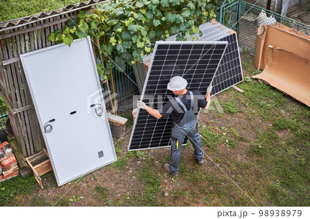 Man technician carrying photovoltaic solar moduls on roof of house. Electrician in helmet installing solar panel system outdoors. Concept of alternative and renewable energy. 98938979