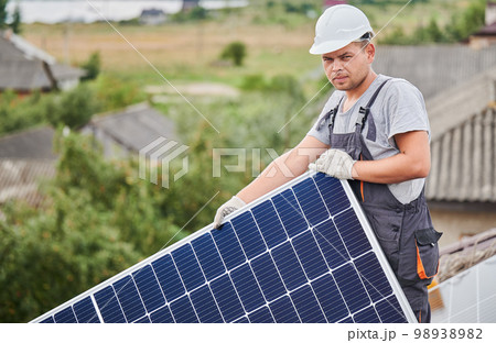 Portrait of man worker mounting photovoltaic solar moduls on roof of house. Electrician in helmet installing solar panel system outdoors. Concept of alternative and renewable energy. Portrait of man worker mounting photovoltaic solar moduls on roof of house. Electrician in helmet installing solar panel system outdoors. Concept of alternative and renewable energy. 98938982