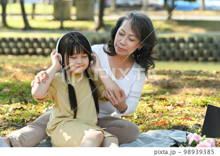 Joyful little girl listening to music in headphones while relaxing with mature grandma in the park on beautiful day Joyful little girl listening to music in headphones while relaxing with mature grandma in the park on beautiful day 98939385