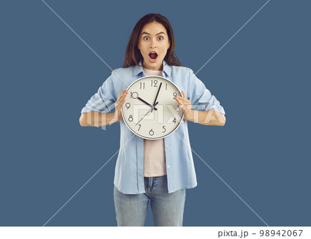 Shocked woman in casual clothes is holding clock in her hands on blue background. Shocked woman in casual clothes is holding clock in her hands on blue background. 98942067