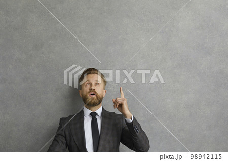 Surprised young businessman looking and pointing up on grey copy space background Surprised young businessman looking and pointing up on grey copy space background 98942115