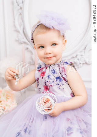 Baby girl elegant dress. A one-year-old girl in a puffy dress and a cute bow poses against the backdrop of a bright room with a dressing table and flowers. 98943922