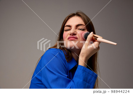 Half-length portrait of young emotional beautiful girl holding blush brush and doing makeup isolated over grey background. Beauty, fashion, style, fun and joy concept Half-length portrait of young emotional beautiful girl holding blush brush and doing makeup isolated over grey background. Beauty, fashion, style, fun and joy concept 98944224