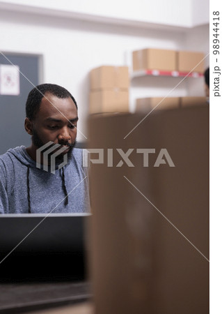 Storehouse manager checking shipment logistics before start preparing packages, putting customer order in carton boxes. African american supervisor working in warehouse delivery department Storehouse manager checking shipment logistics before start preparing packages, putting customer order in carton boxes. African american supervisor working in warehouse delivery department 98944418