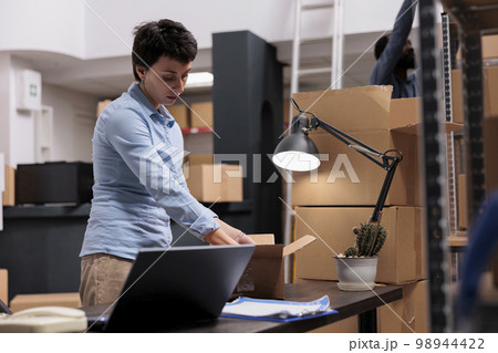 Caucasian manager preparing customers orders putting trendy clothes in carton boxes while looking at cargo stock on laptop computer. Workers working in delivery department in warehouse Caucasian manager preparing customers orders putting trendy clothes in carton boxes while looking at cargo stock on laptop computer. Workers working in delivery department in warehouse 98944422