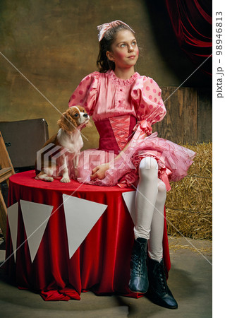 Dreaming. Beautiful little girl in festive pink dress posing with cute dog over dark vintage circus background. Holidays, dreams, art, fashion, vintage style 98946813
