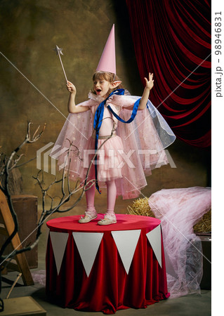 Little performer. Beautiful cute girl, child in image of magical elf posing in pink dress over vintage circus background. Making magic Little performer. Beautiful cute girl, child in image of magical elf posing in pink dress over vintage circus background. Making magic 98946831