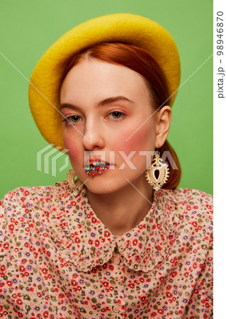Beautiful young girl in cute outfit with yellow beret, blushing cheeks and candy toppings on lips over green background. Food pop art photography. Complementary colors 98946870