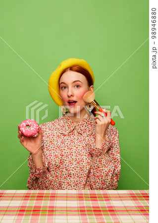 Cute looking. Beautiful girl with blushing cheeks eating pink donut over green background. Countryside style. Food pop art photography. Complementary colors Cute looking. Beautiful girl with blushing cheeks eating pink donut over green background. Countryside style. Food pop art photography. Complementary colors 98946880
