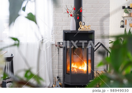 Valentine decor near fireplace stove with fire and firewood. Cozy home hearth in interior with potted plans, valentines day in family love nest. Valentine decor near fireplace stove with fire and firewood. Cozy home hearth in interior with potted plans, valentines day in family love nest. 98949030
