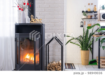 Valentine decor near fireplace stove with fire and firewood. Cozy home hearth in interior with potted plans, valentines day in family love nest. Valentine decor near fireplace stove with fire and firewood. Cozy home hearth in interior with potted plans, valentines day in family love nest. 98949036