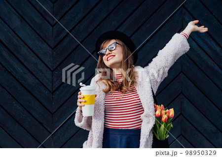 Happy, emotional young fashion woman with reusable coffee cup and net bag with fresh tulip flowers inside on the black wooden background. Urban city street fashion. Spring mood. Selective focus 98950329