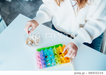 Woman's hands sorting pills into pills organizer. Close up medical pill box with daily doses of medicines. Antioxidant diet vitamin supplements for health and beauty care, biohacking concept. Woman's hands sorting pills into pills organizer. Close up medical pill box with daily doses of medicines. Antioxidant diet vitamin supplements for health and beauty care, biohacking concept. 98950334