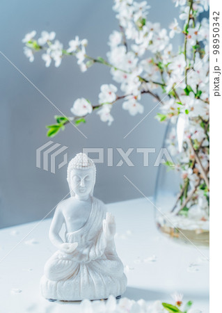 Decorative white Buddha statuette with blooming tree branches in the vase on the light background. Meditation and relaxation ritual. Buddha birthday. Vertical card. Selective focus 98950342