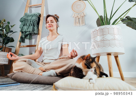 Young healthy woman practicing meditation at home, wearing headset, sitting in lotus position on floor cushion in modern Scandi interior home. Meditating, relaxed, eyes closed. Selective focus. Young healthy woman practicing meditation at home, wearing headset, sitting in lotus position on floor cushion in modern Scandi interior home. Meditating, relaxed, eyes closed. Selective focus. 98950343