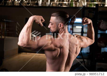 A handsome bearded male athlete with a muscular body poses and shows his biceps and back in the gym 98951712