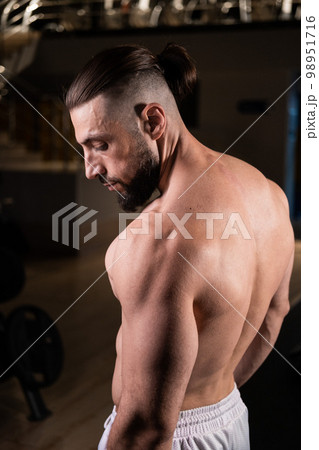 A handsome bearded male athlete with a muscular body poses in the gym in white pants. Active lifestyle, sports A handsome bearded male athlete with a muscular body poses in the gym in white pants. Active lifestyle, sports 98951716