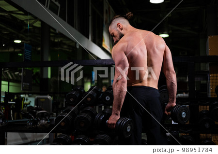 A brutal muscular bearded man stands with his back and holds a dumbbell in the gym. Active lifestyle, sports A brutal muscular bearded man stands with his back and holds a dumbbell in the gym. Active lifestyle, sports 98951784