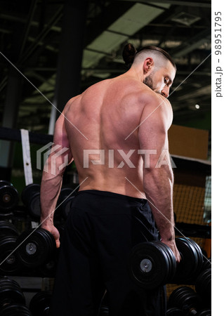 A brutal muscular bearded man stands with his back and holds a dumbbell in the gym. Active lifestyle, sports 98951795