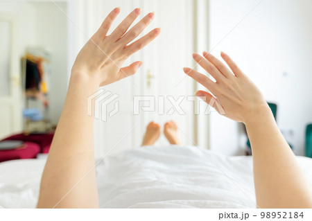 Woman lies in bed and stretches after waking up. Beautiful white room in the background. Hands close up Woman lies in bed and stretches after waking up. Beautiful white room in the background. Hands close up 98952184