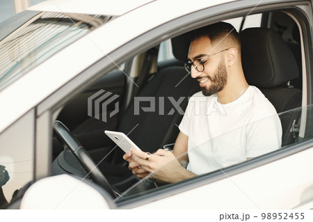 Handsome indian young man looking at his smart phone while sitting in the car. Man wearing white t-shirt and glasses. Male driver sitting on car and hand holding smartphone to use online application. 98952455
