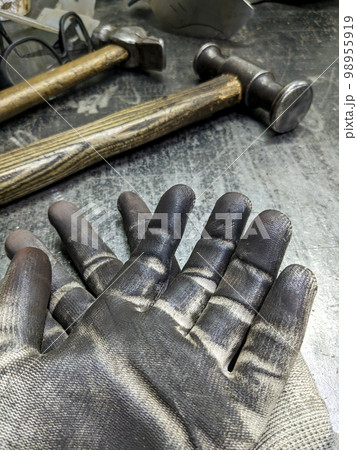 Black blacksmith work gloves against a sledgehammer background. Hard men's work as a blacksmith. Black blacksmith work gloves against a sledgehammer background. Hard men's work as a blacksmith. 98955919