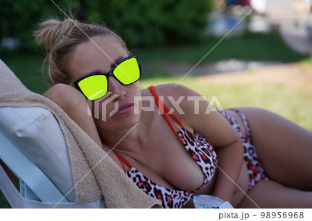 A curvy-breasted blonde in a colorful swimsuit and chroma key sunglasses lies on her side on the beach and stares at you intently. Portrait of a young woman on the beach. 98956968