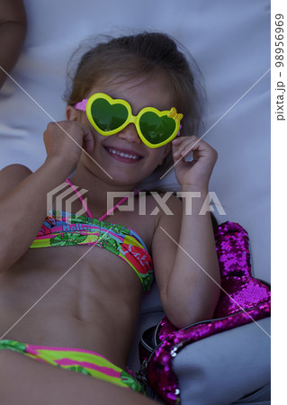 Little girl in funny yellow sunglasses with green glasses in form of hearts laughs merrily. Child on the beach in swimsuit is having fun. A cheerful white girl. Selective focus 98956969