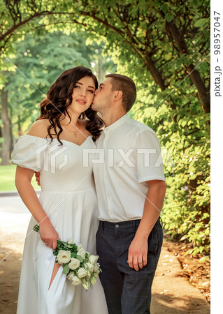 Beautiful white young couple strolls in garden. Black-haired girl in festive dress and with a bouquet of white flowers. Man in white shirt gently kisses his companion. Portrait of young couple 98957047