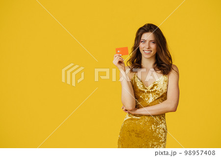 Positive woman in dress holding credit card and smiling at camera, shopping contactless, yellow background, free space Positive woman in dress holding credit card and smiling at camera, shopping contactless, yellow background, free space 98957408