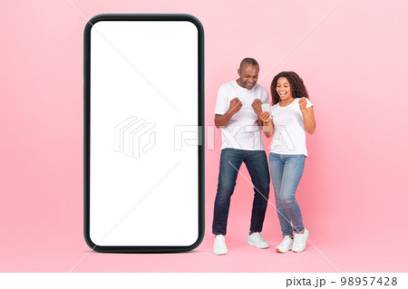 Excited african american spouses standing near huge smartphone with empty screen, using phone and shaking clenched fists Excited african american spouses standing near huge smartphone with empty screen, using phone and shaking clenched fists 98957428