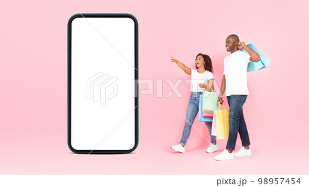 Excited black couple with shopping bags walking near giant smartphone with empty screen, pink background, mockup 98957454