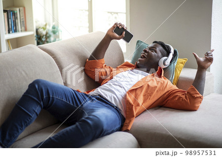 Happy black guy lying on sofa, singing songs, using phone Happy black guy lying on sofa, singing songs, using phone 98957531