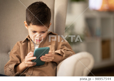 Busy serious pretty caucasian little child with smartphone sits in armchair, watches videos in living room 98957544