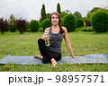 Young woman in comfortable sportwear sitting left twist on yoga mat with a water bottle relaxed after workout outside in the green park. 98957571