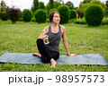 Young woman in comfortable sportwear sitting left twist on yoga mat with a water bottle relaxed after workout outside in the green park.  98957573