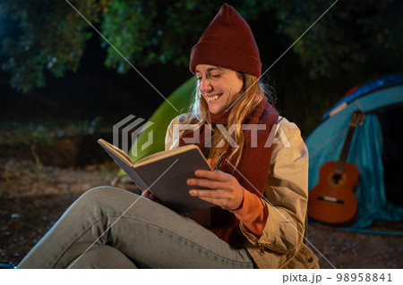 Girl reading a book at night camping. Night scene next to camping fire. Relaxing in quiet nature Girl reading a book at night camping. Night scene next to camping fire. Relaxing in quiet nature 98958841