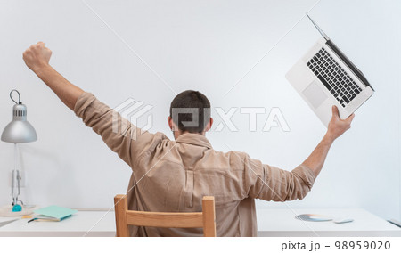 Unrecognizable young man with laptop celebrating with raised arms success at work in home office. Unrecognizable young man with laptop celebrating with raised arms success at work in home office. 98959020