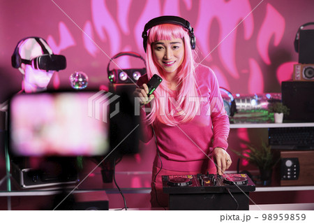 Asian artist playing techno song at mixer console while recording music session using professional camera, preparing to perform in night club. Dj with pink hair using audio equipment 98959859