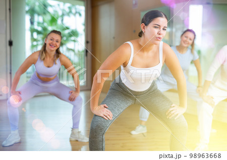 Cheerful young mature female dancers in sportswear dancing with legs wide apart during exercising with coach in gym Cheerful young mature female dancers in sportswear dancing with legs wide apart during exercising with coach in gym 98963668