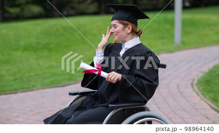 A caucasian woman in a wheelchair holds her diploma and cries with joy outdoors. A caucasian woman in a wheelchair holds her diploma and cries with joy outdoors. 98964140